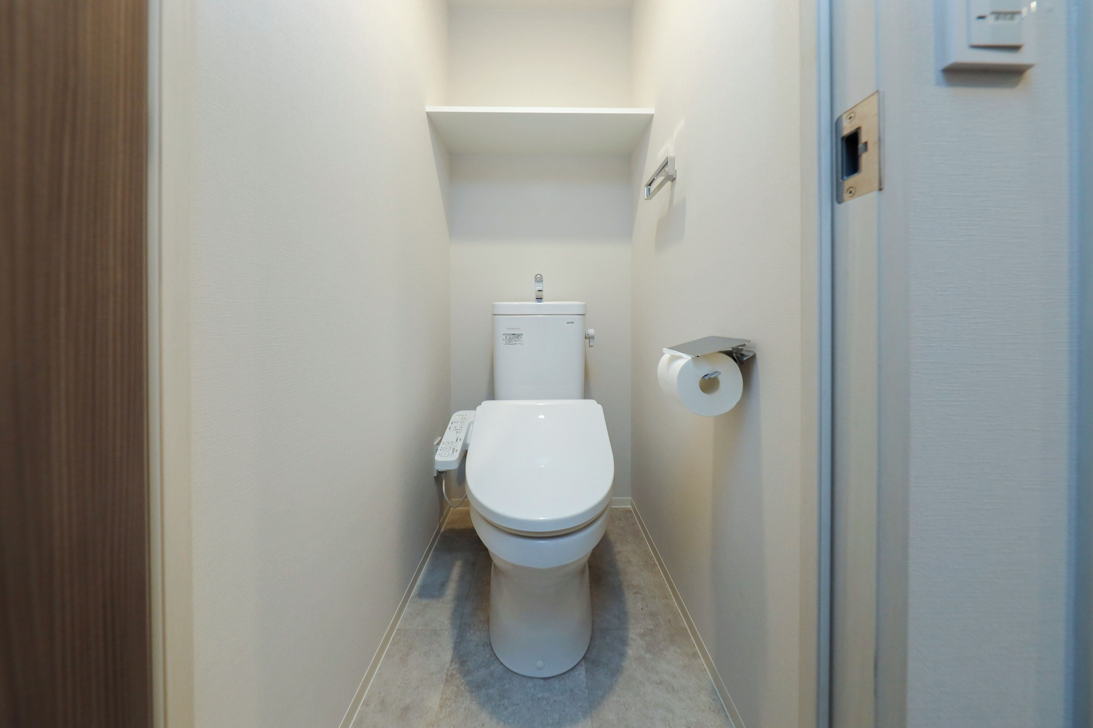 A compact bathroom with a white toilet, a small shelf above the toilet, and a flush handle on the wall.