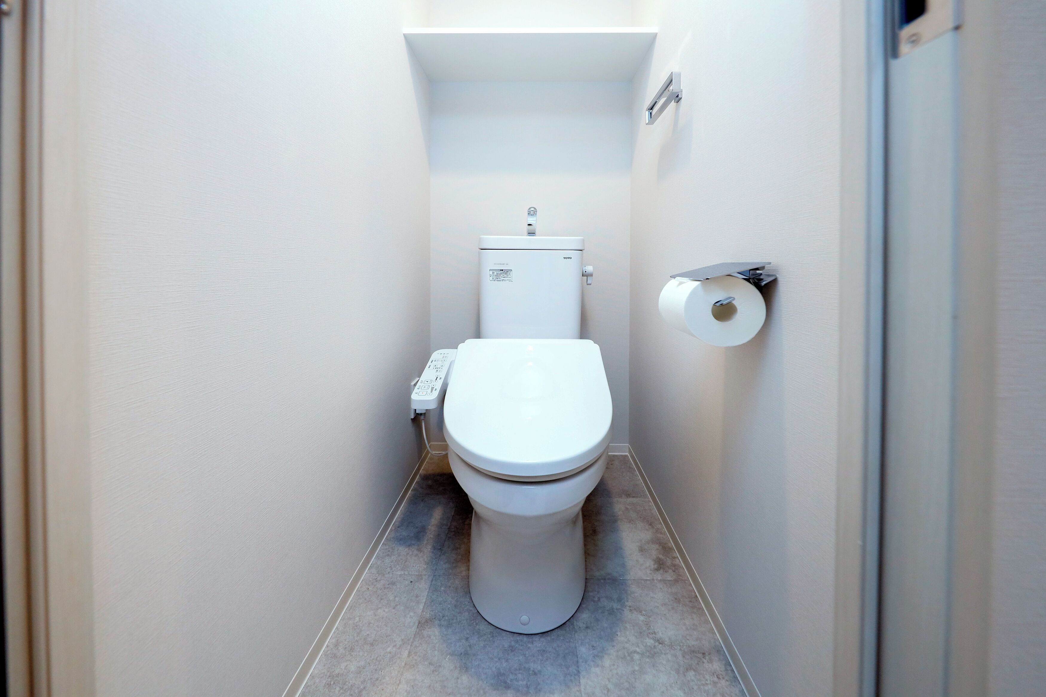 A small, narrow bathroom with a white toilet, a toilet paper holder, and a wall-mounted flush handle.