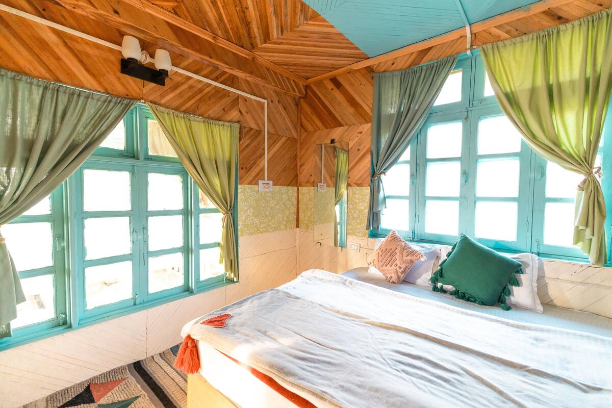 Ein Zimmer mit Holzboden, großen Fenstern mit grünen Vorhängen, einem Bett mit weißer Bettwäsche und einem gemusterten Teppich.