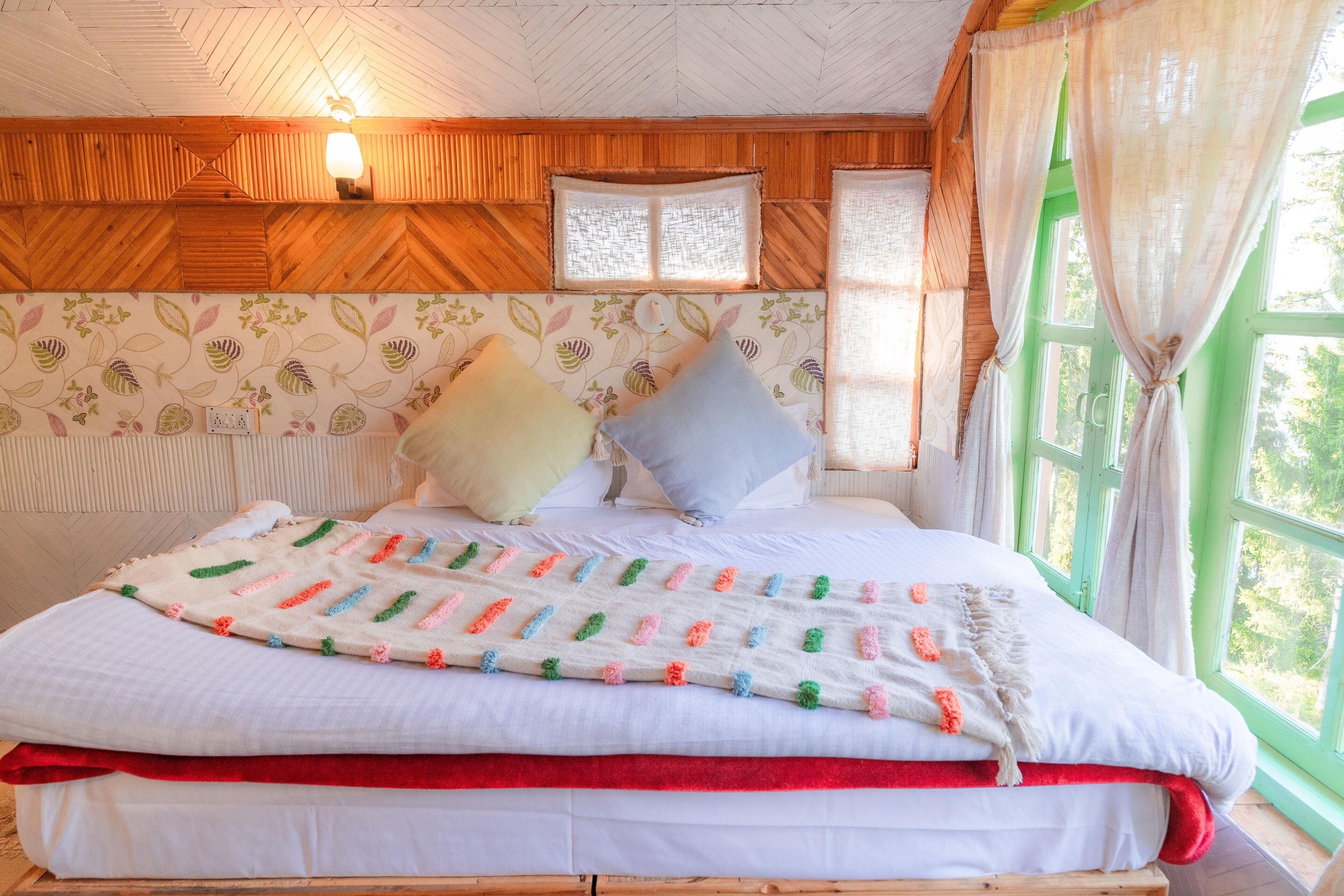 A bed with a colorful blanket, pillows, and a red bed skirt in a room with wooden walls, a window with curtains, and a wall-mounted light fixture.