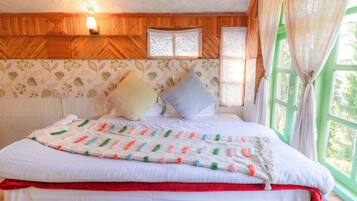 A bed with a colorful blanket, pillows, and a red bed skirt in a room with wooden walls, a window with curtains, and a wall-mounted light fixture.