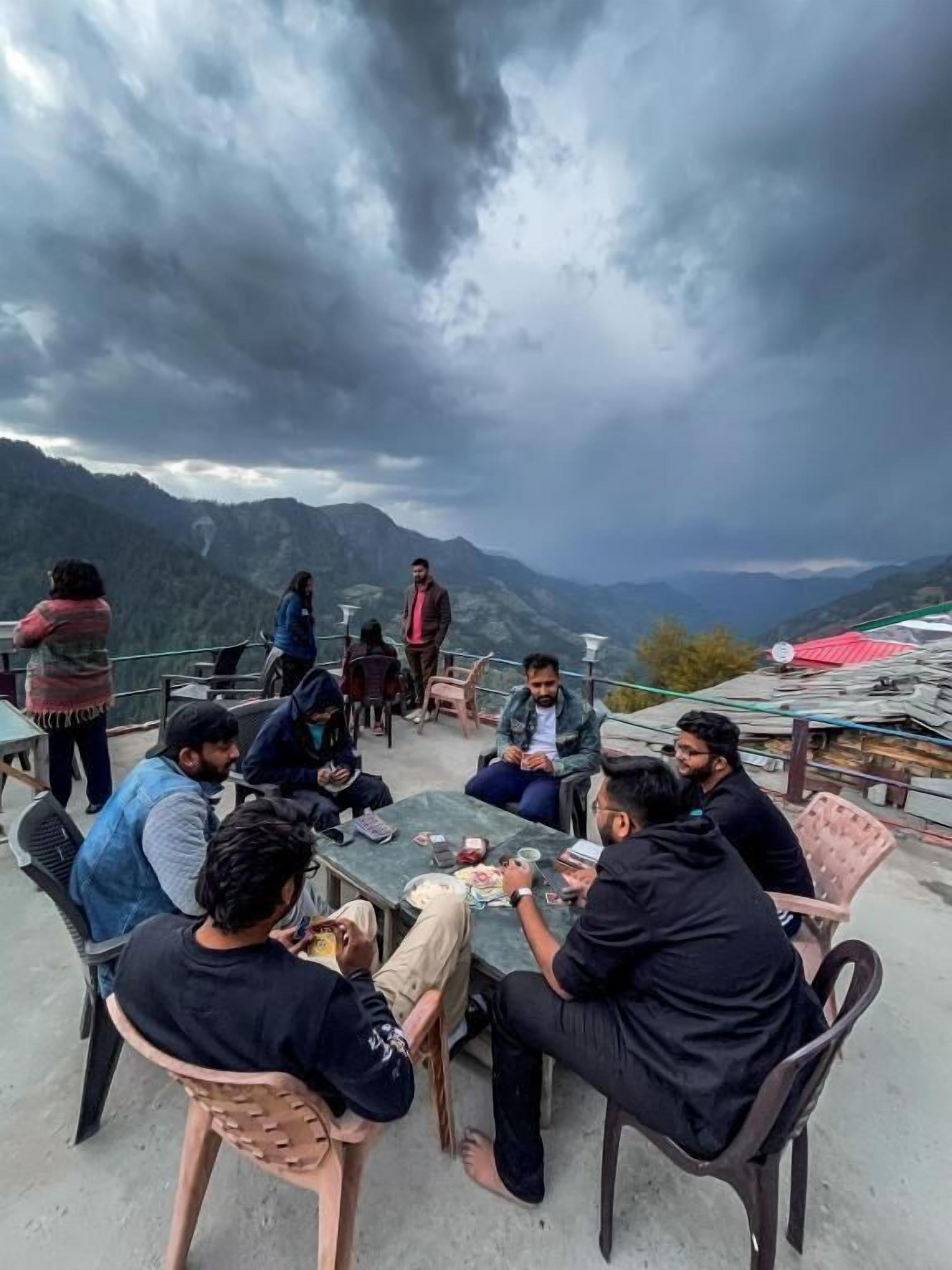 Un grupo de personas sentadas alrededor de una mesa al aire libre, con montañas al fondo.