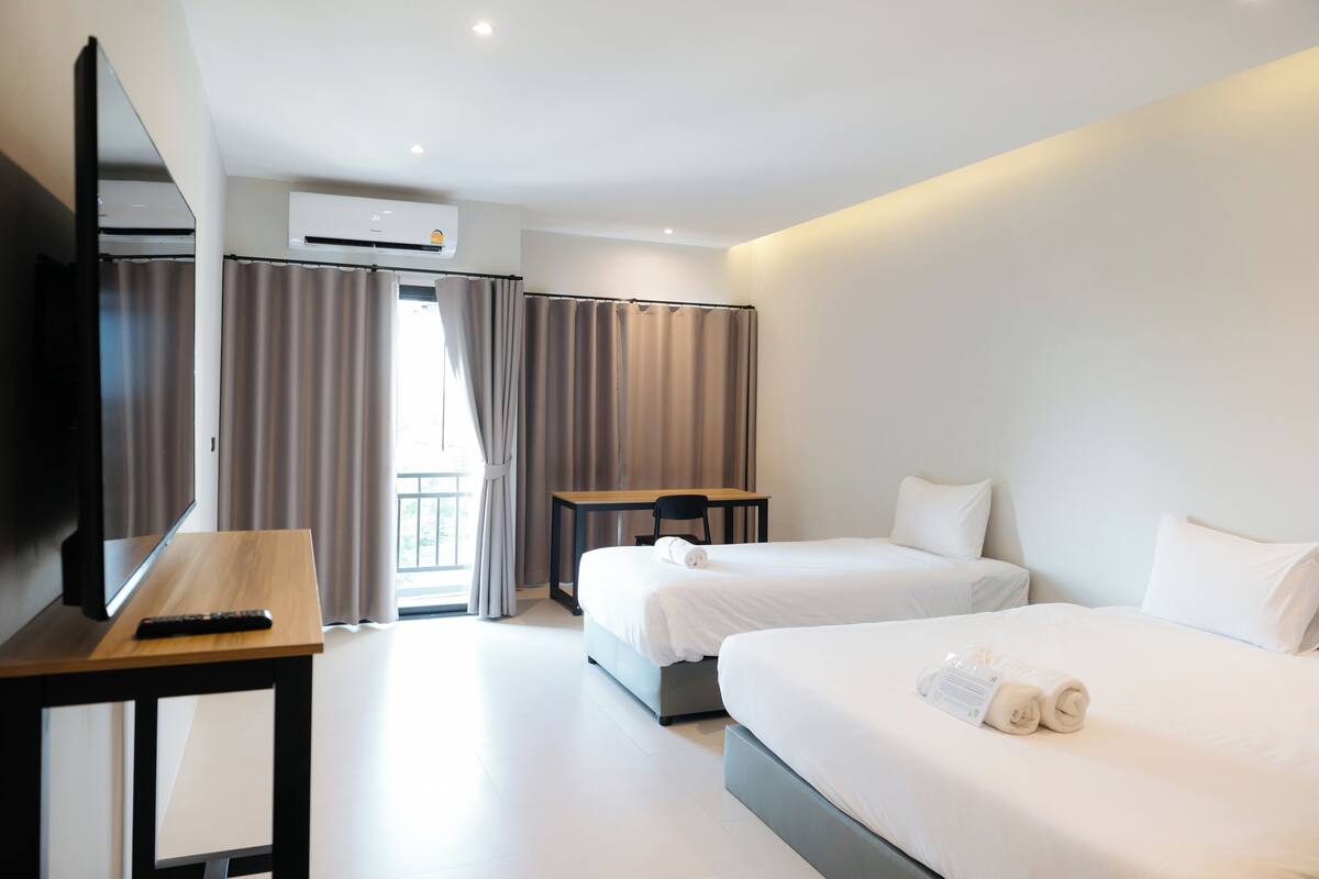 A hotel room with two beds, a desk, a TV, and an air conditioning unit.