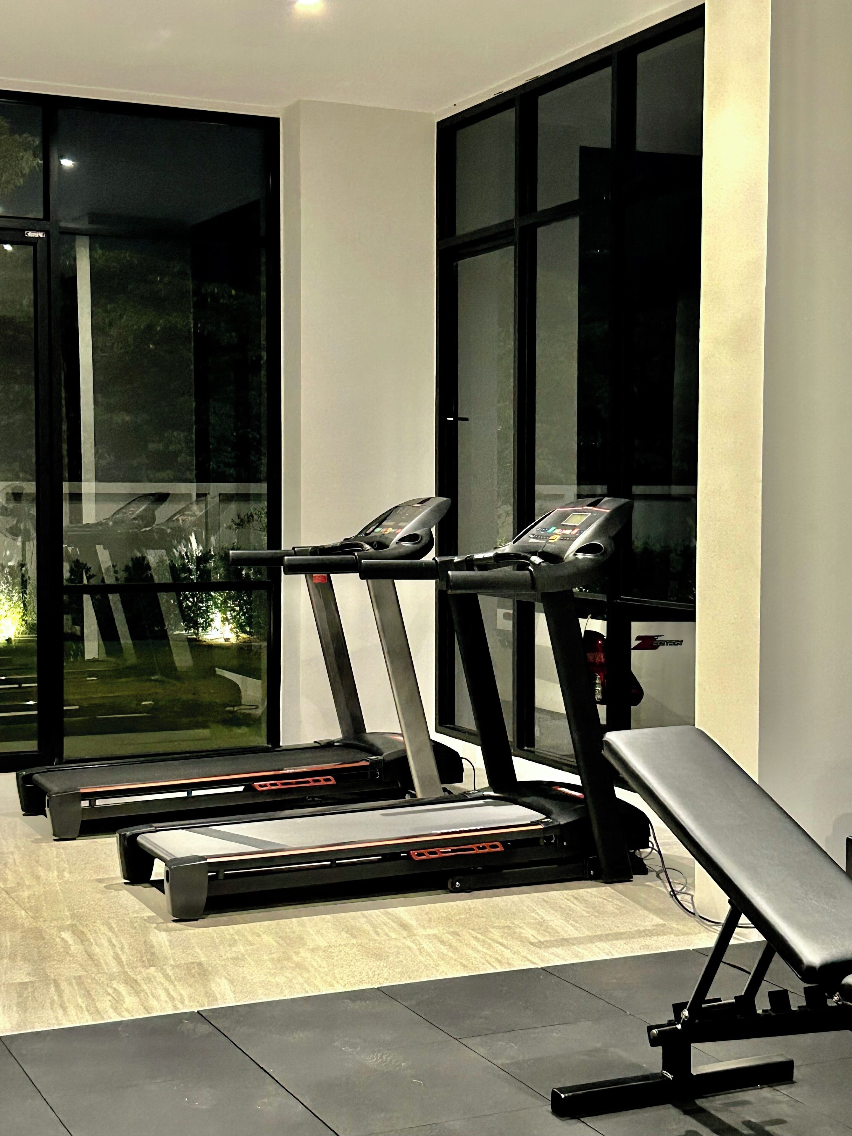 Et fitnesscenter med løbebånd og bænk, store vinduer og udsigt til det fri.
