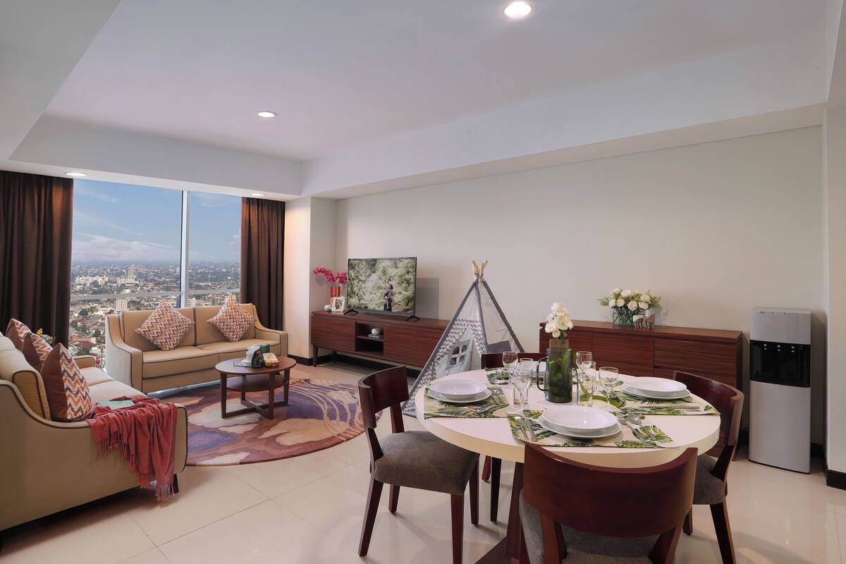 A modern living room with a dining area, large windows, a sofa, a coffee table, and a TV.
