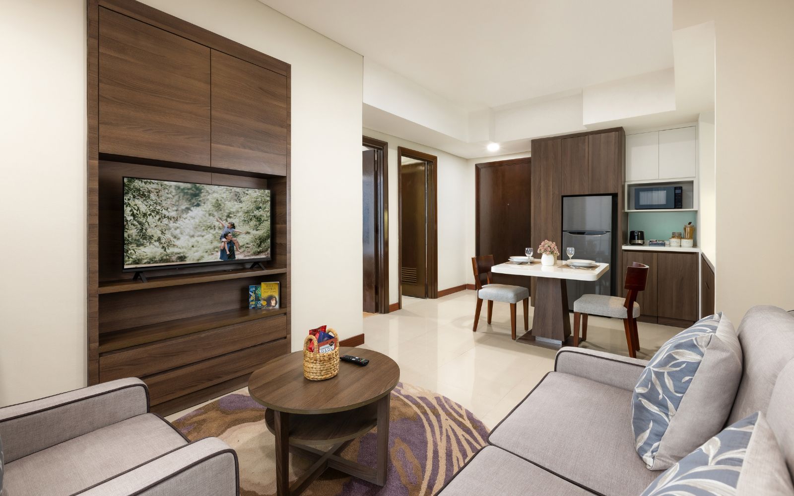 A modern living room with a flat-screen TV, a dining area with a table and chairs, and a kitchen area with a refrigerator.