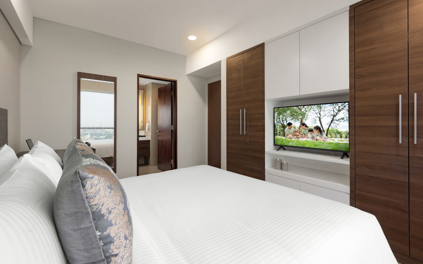 Une chambre moderne avec un grand lit, un téléviseur fixé sur un meuble en bois et une vue sur un parc, visible par la fenêtre.