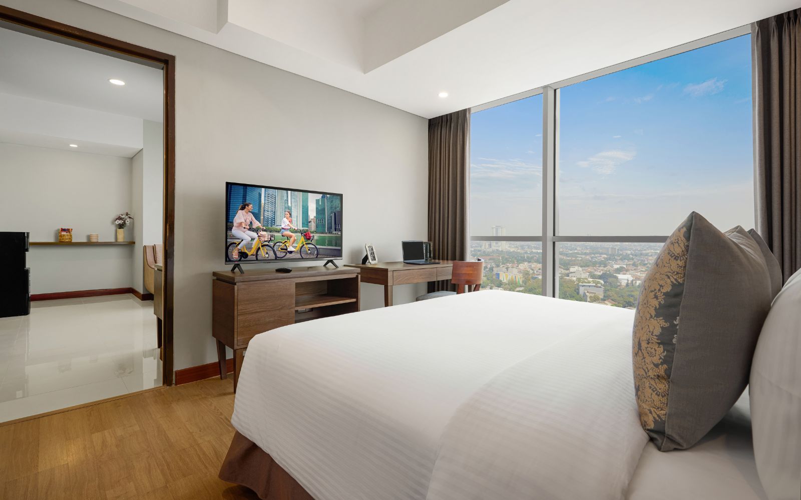 Una habitación de hotel con una cama grande, un escritorio con una computadora portátil, un televisor y vistas a un paisaje urbano.