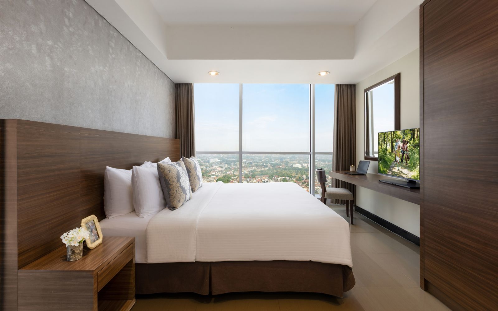 A modern hotel room with a large bed, a desk with a laptop, a television, and a city view.