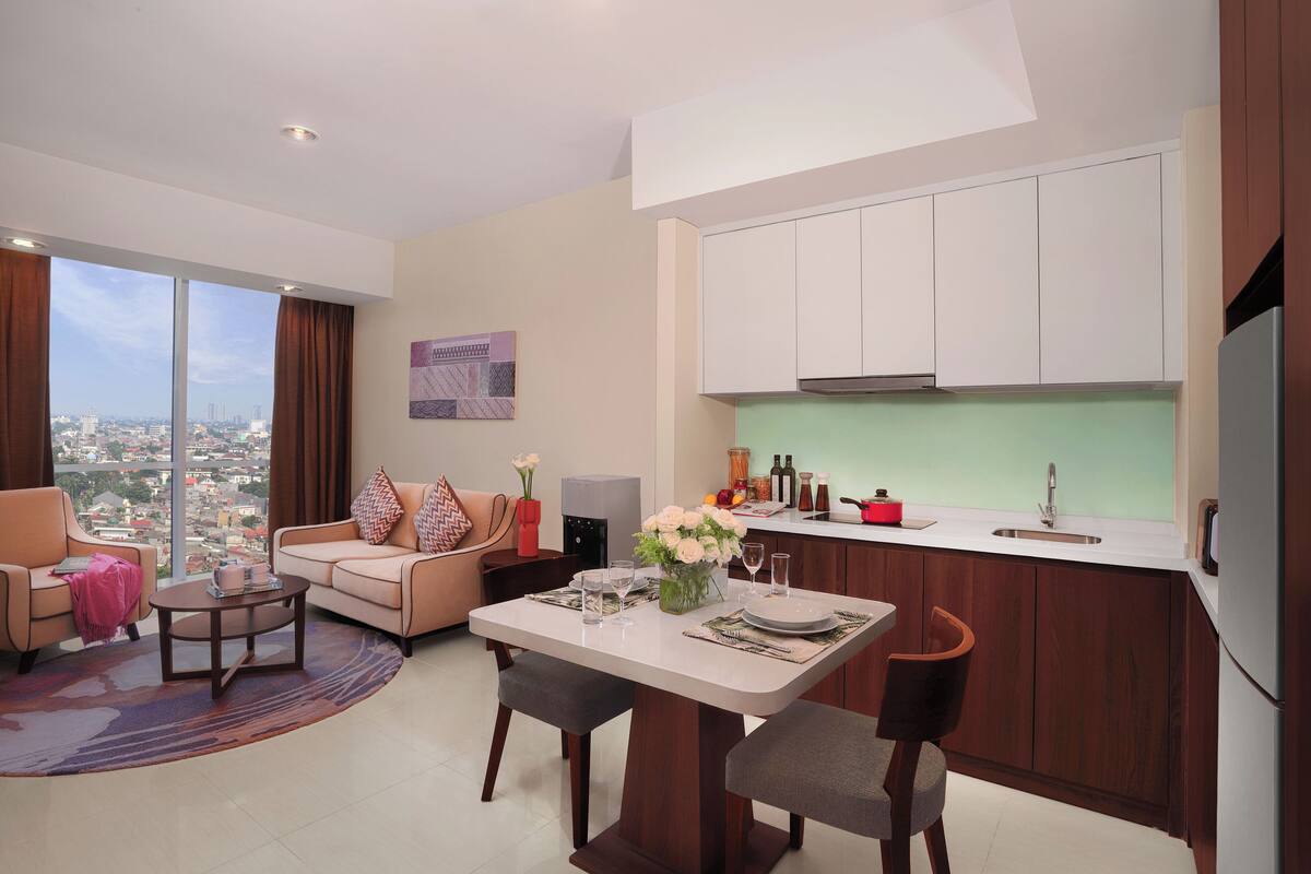 A modern living room with a dining area, a kitchen with white cabinets, and a large window offering a city view.