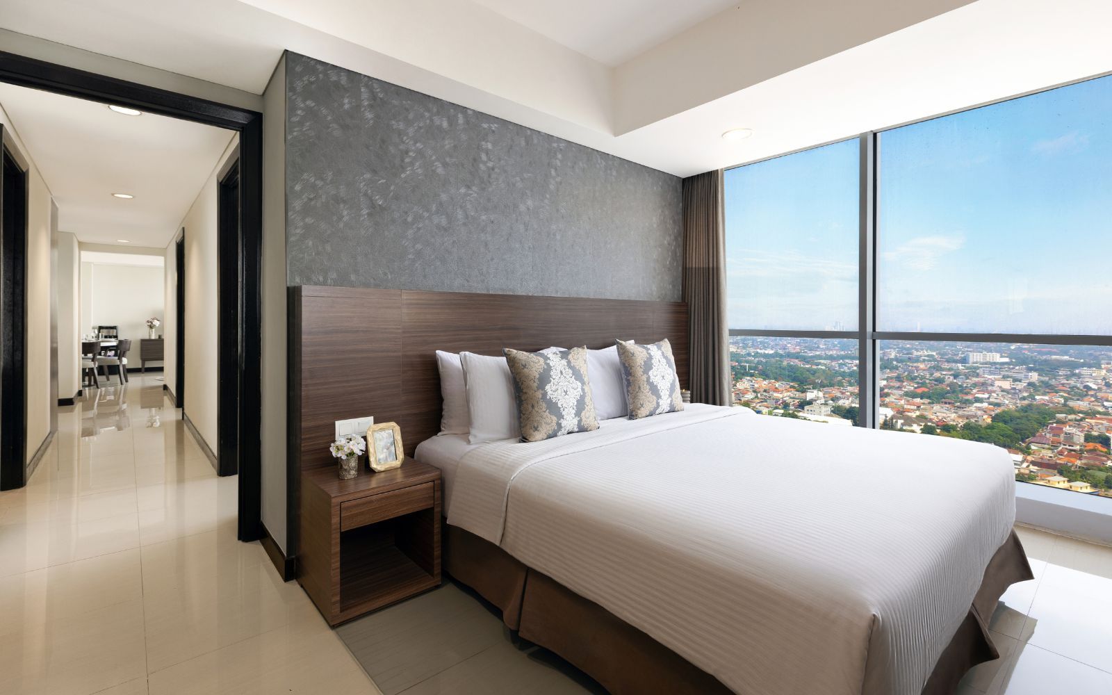 A modern hotel room with a large bed, a nightstand, a view of the city, and a separate dining area.