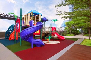 A playground with slides and climbing structures, surrounded by greenery and a building in the background.