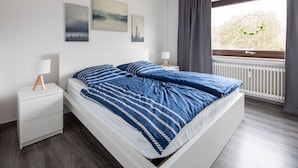 A modern bedroom with a white bed, blue striped bedding, and a view of a balcony with a railing and greenery. - Ferienwohnung Watt, Modern und Liebevoll Eingerichtet, Wlan, Smart TV (Wilhelmshaven)