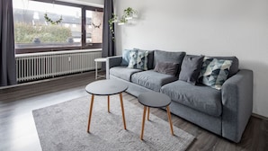 A modern living room with a grey sofa, two small round tables, a grey rug, a radiator, and a window with curtains. - Ferienwohnung Watt, Modern und Liebevoll Eingerichtet, Wlan, Smart TV (Wilhelmshaven)