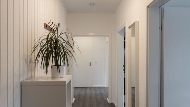 A hallway with a wooden floor, white walls, a potted plant, a coat rack, and a door at the end. - Ferienwohnung Watt, Modern und Liebevoll Eingerichtet, Wlan, Smart TV (Wilhelmshaven)