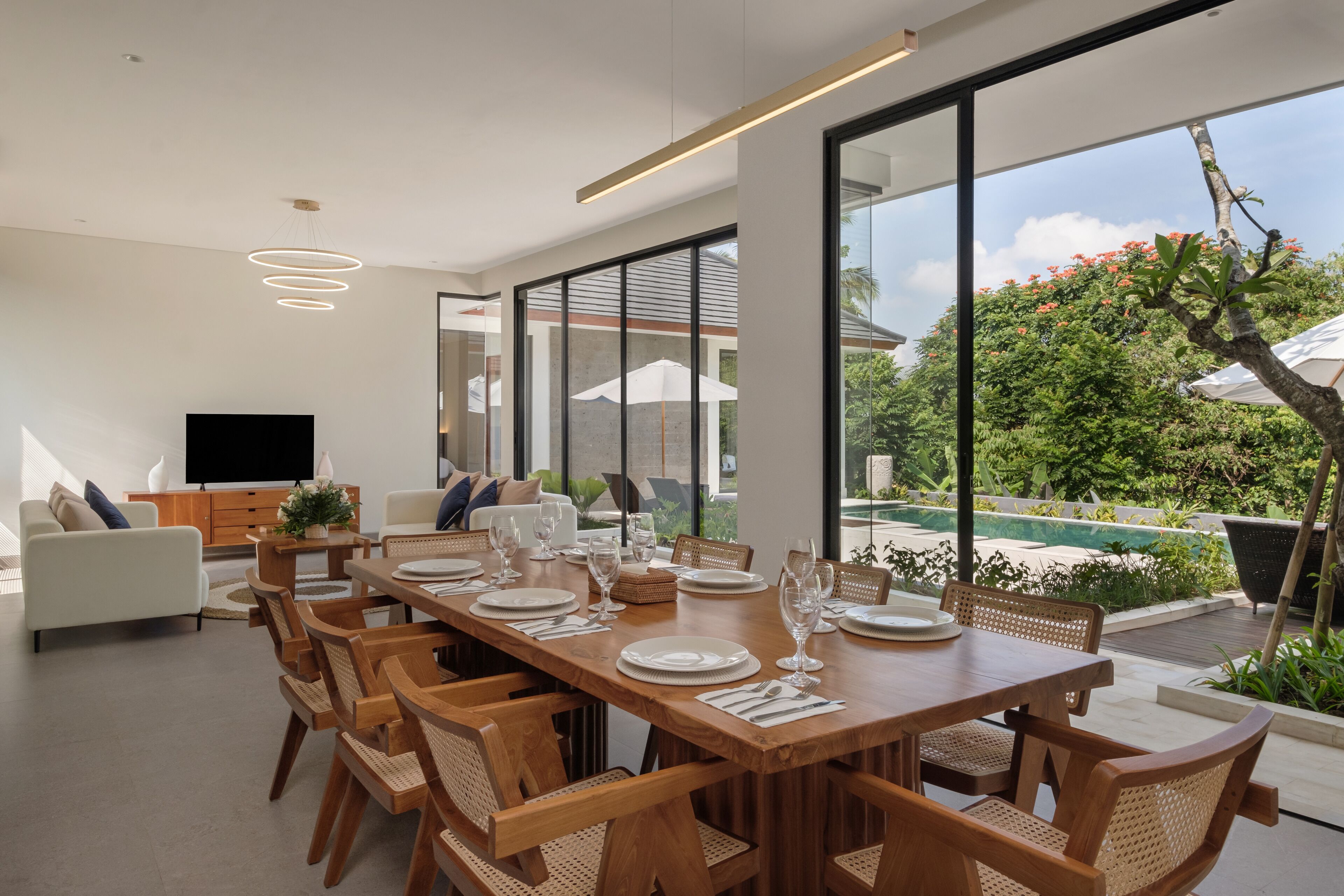 Un comedor moderno con una mesa de madera dispuesta para seis personas, rodeado de sillas de mimbre. Un ventanal ofrece vistas a una piscina y abundante vegetación.