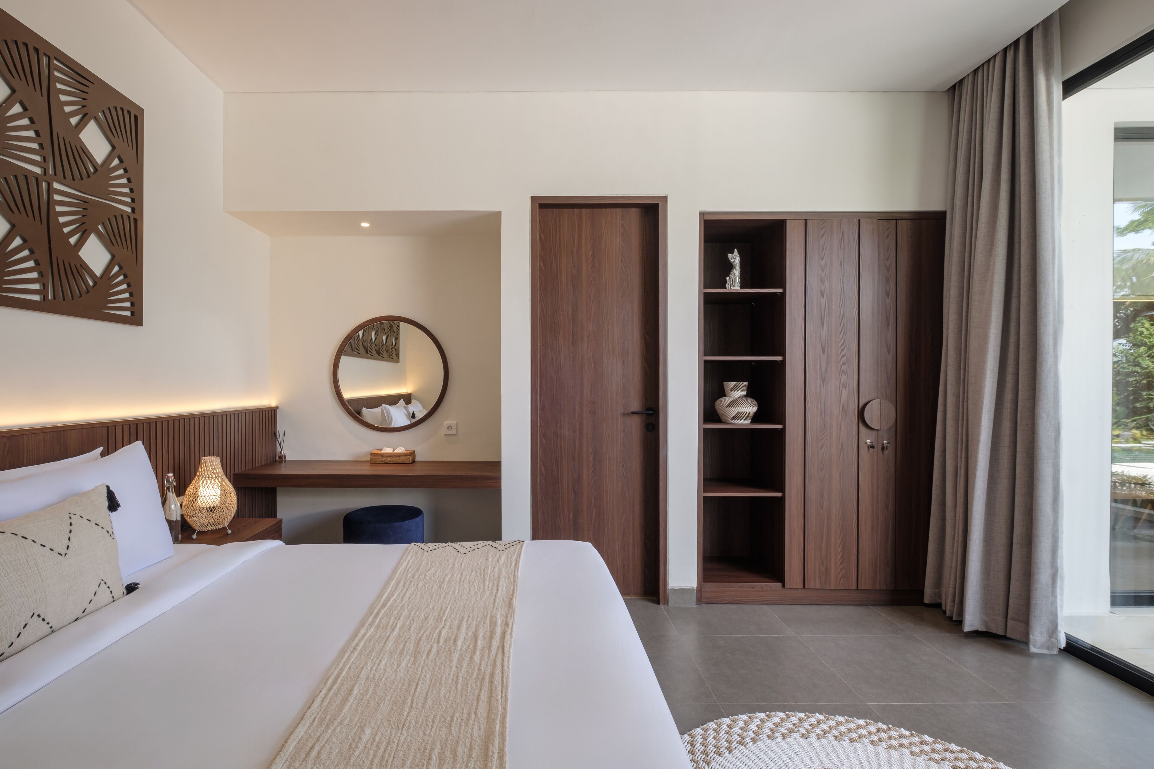 A modern hotel room with a large bed, a wooden wardrobe, a desk with a lamp, and a large window with curtains.