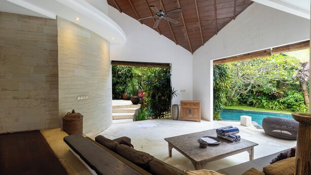 A modern interior with a stone wall, a wooden ceiling, a ceiling fan, a sofa, a coffee table, and a view of a swimming pool and greenery outside.