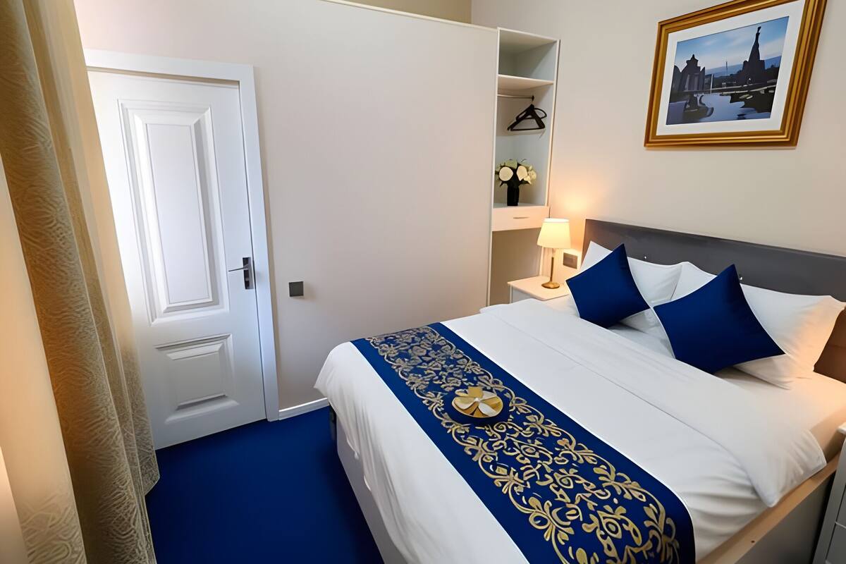 Habitación de hotel con una cama, una mesita de noche, una lámpara, un cuadro en la pared y una puerta.