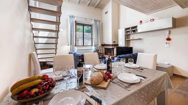 A dining area with a table set for four, including plates, cutlery, and a loaf of bread. A bowl of fruit is on the table, and there is a bottle of wine. The room has a wooden ceiling and a window with curtains. - Trekking Home La Via degli Dei Toscana (Vaglia)