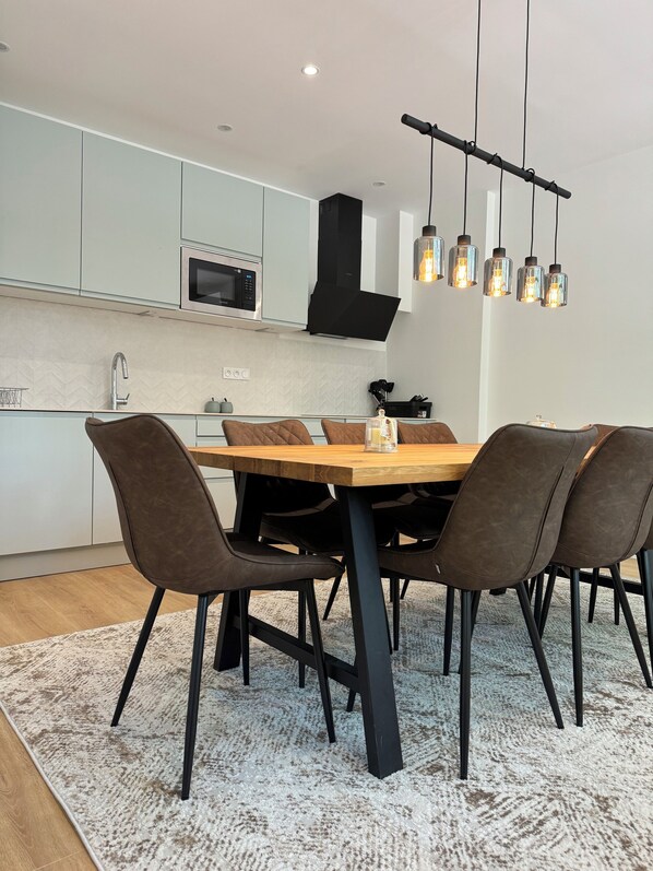 A modern kitchen with white cabinets, a microwave, and a black range hood. There is a dining table with six chairs and a hanging light fixture above.