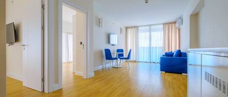 Un salón espacioso con piso de madera, un sofá azul, un televisor montado en la pared y una zona de comedor con sillas azules.
