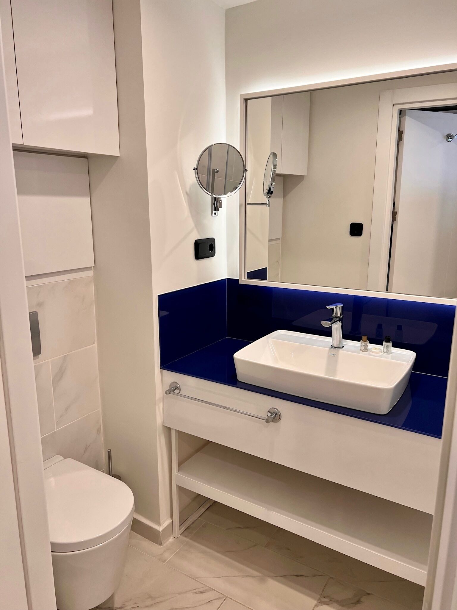 A modern bathroom with a white toilet, a blue vanity with a white sink, and a large mirror.