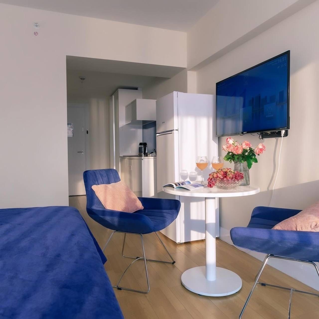 A modern room with two blue chairs, a white table with a vase of flowers, and a flat-screen TV mounted on the wall.