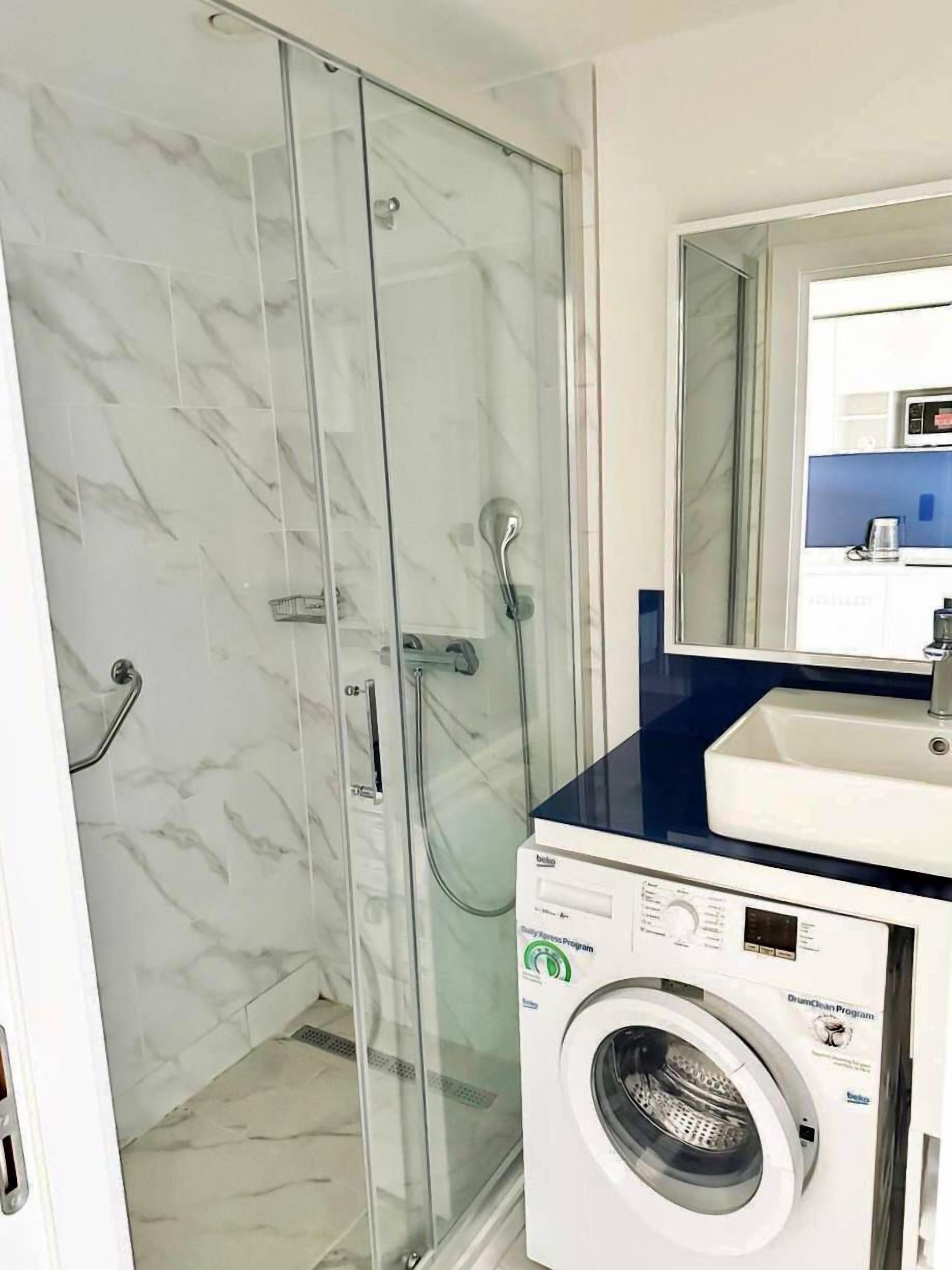 Een badkamer met een douche in een glazen cabine, een witte wasmachine en een wastafel met een blauw aanrechtblad.