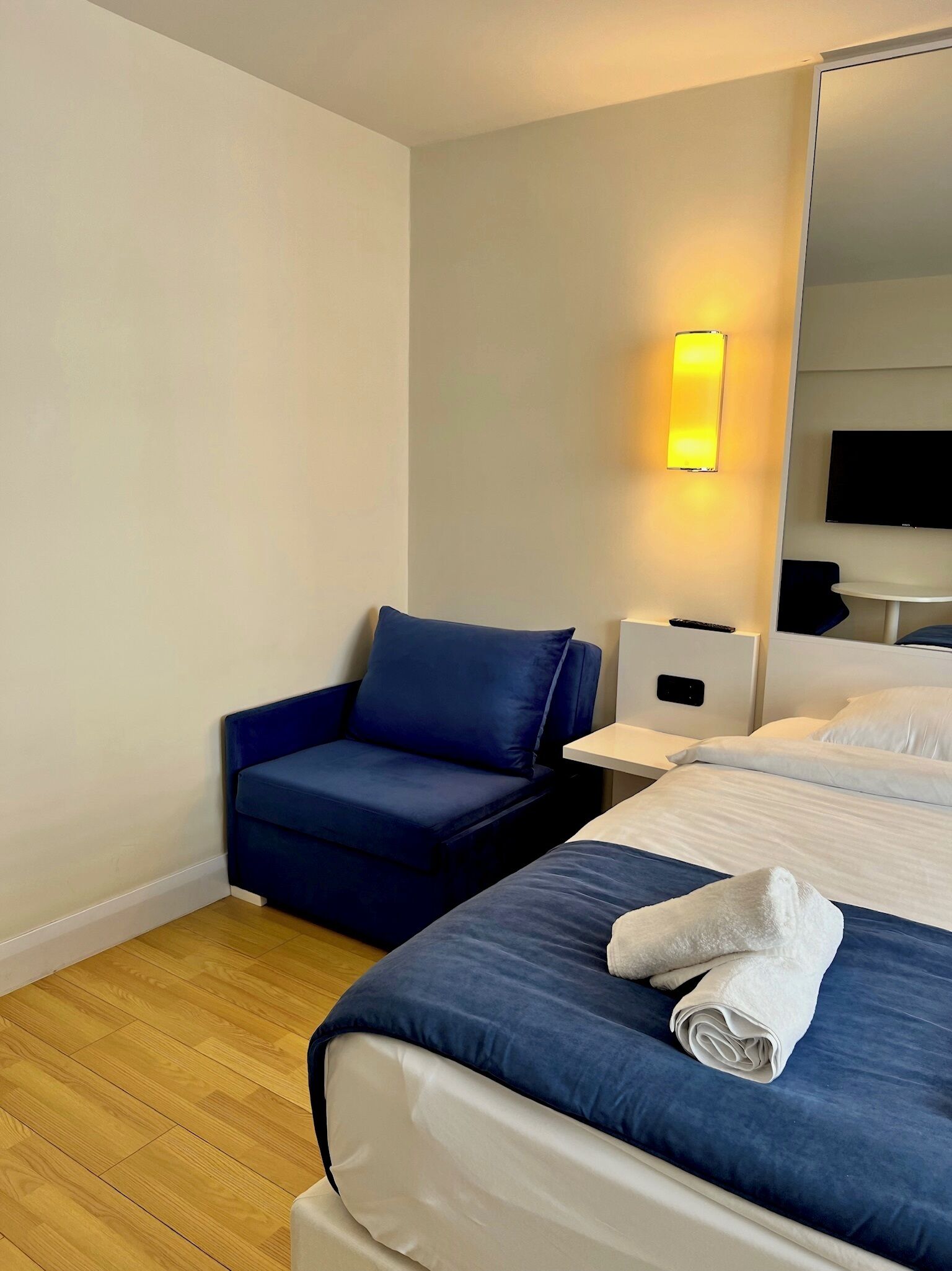 A hotel room with a bed, a blue armchair, a bedside table, a lamp, and a flat-screen TV mounted on the wall.