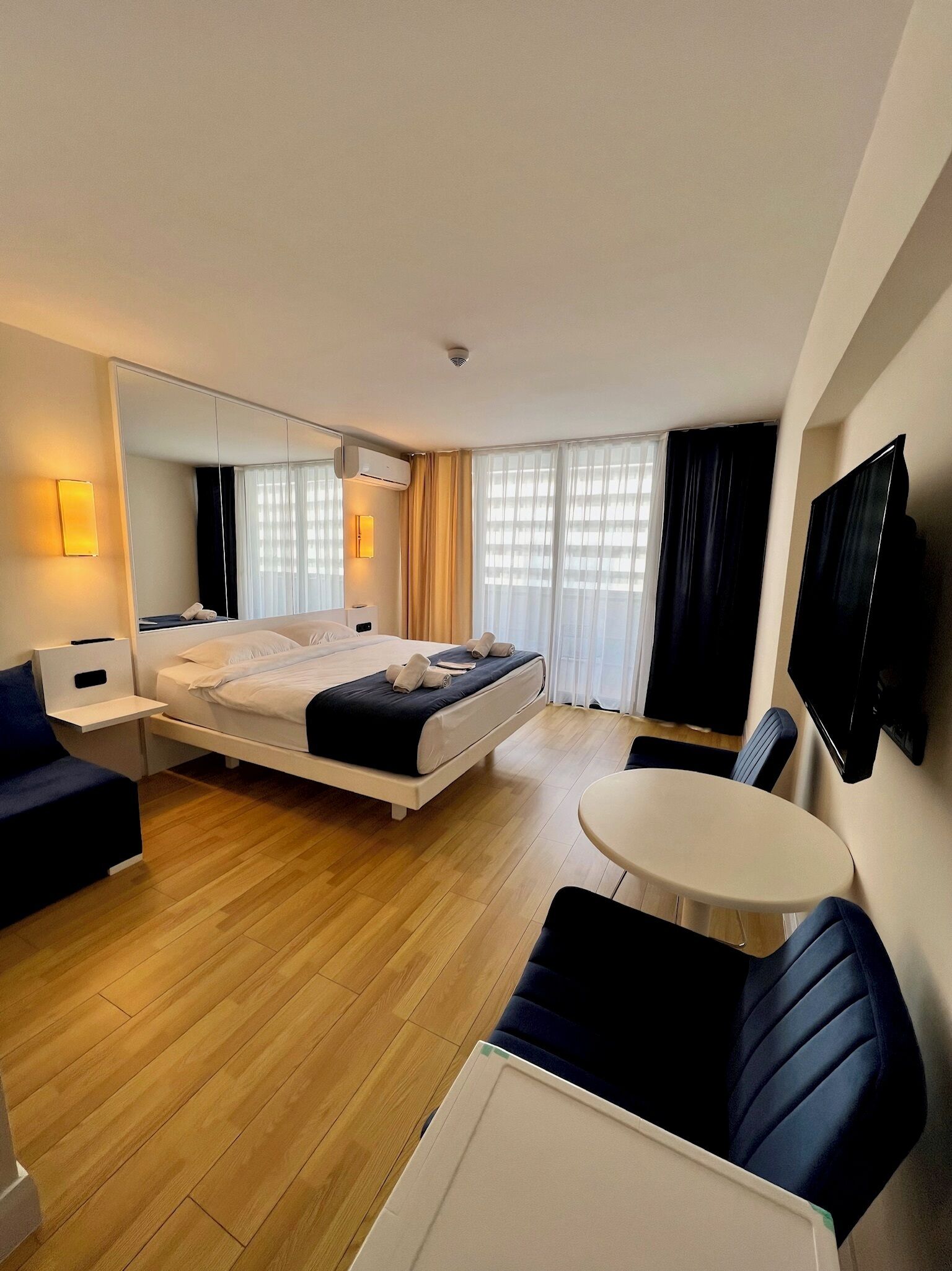 A modern hotel room with a bed, a sofa, a small table, a TV, and a large mirror.