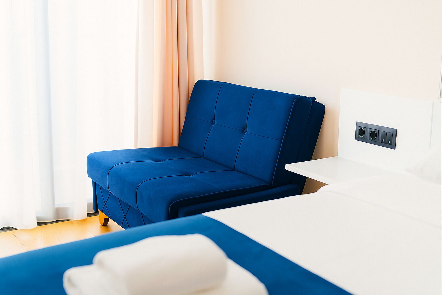 A modern hotel room with a blue sofa, a white bedside table, and a wall-mounted control panel.