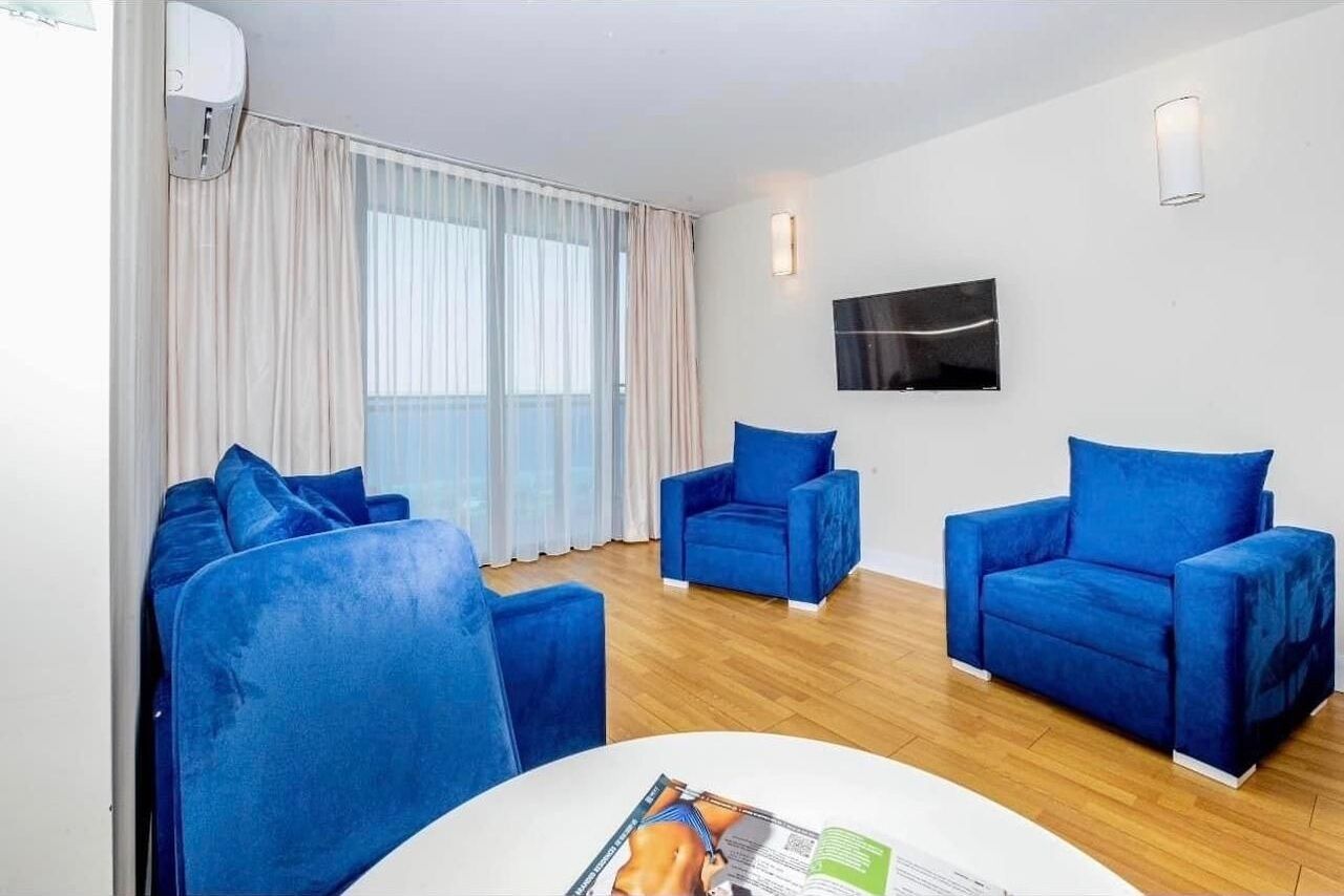 Quarto moderno com piso de madeira, uma mesa redonda e três poltronas azuis. Possui uma janela grande com cortinas, uma televisão de tela plana na parede e um ar-condicionado também na parede.