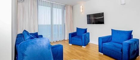 Quarto moderno com piso de madeira, uma mesa redonda e três poltronas azuis. Possui uma janela grande com cortinas, uma televisão de tela plana na parede e um ar-condicionado também na parede.