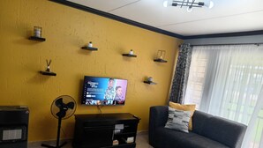 A living room with a yellow wall, a flat-screen TV, a black sofa, a black entertainment unit, and a floor fan.