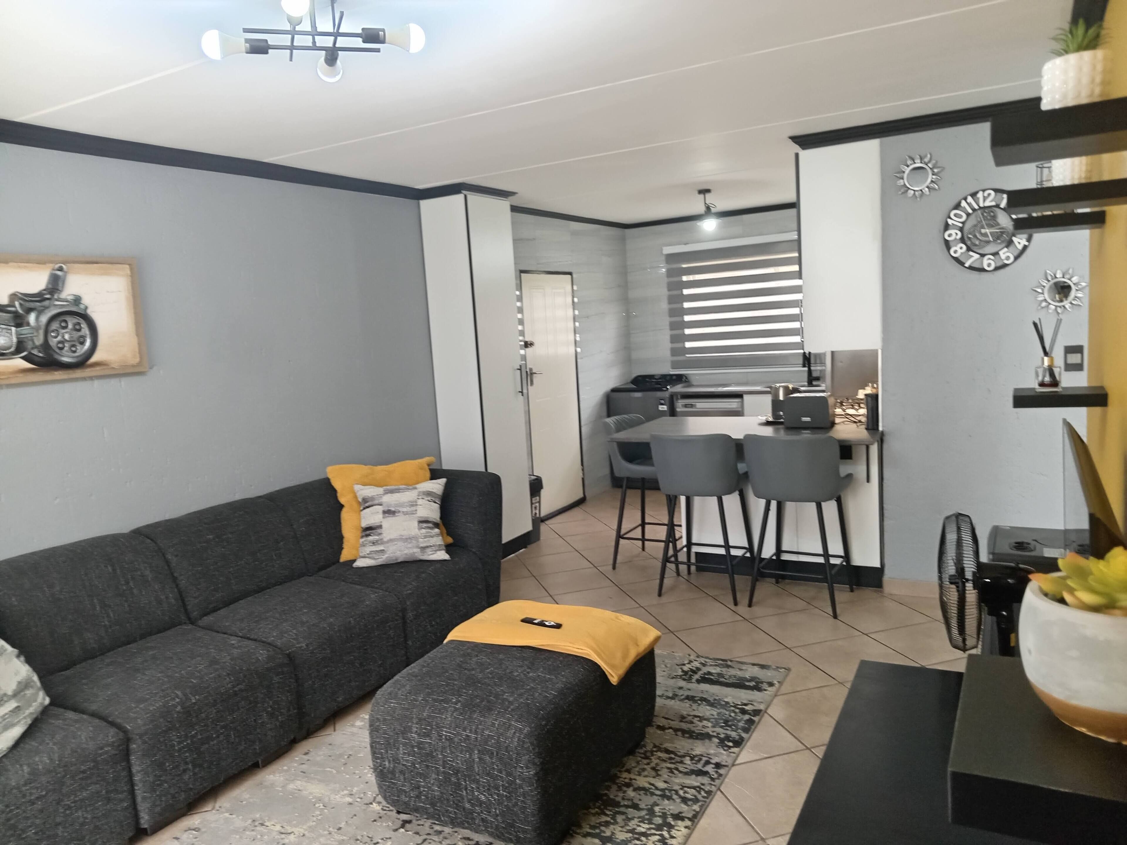 Ein modernes Wohnzimmer mit grauem Sofa, Hocker und einem kleinen Tisch mit Pflanze. Es gibt eine Küchenzeile mit Esstisch und Stühlen sowie einen Deckenventilator.