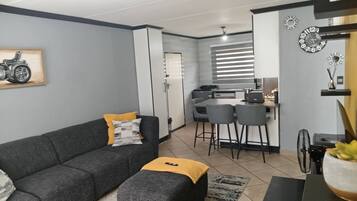 Ein modernes Wohnzimmer mit grauem Sofa, Hocker und einem kleinen Tisch mit Pflanze. Es gibt eine Küchenzeile mit Esstisch und Stühlen sowie einen Deckenventilator.