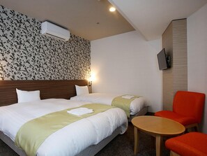 Hotellihuone, jossa on kaksi sänkyä, pieni pöytä, tuoli, ilmastointilaite ja seinälle asennettu televisio.