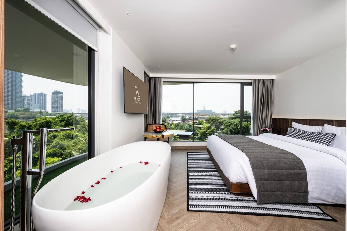 Quarto de hotel moderno com uma cama grande, banheira com pétalas de rosa, vista para a cidade e uma pequena mesa com frutas.