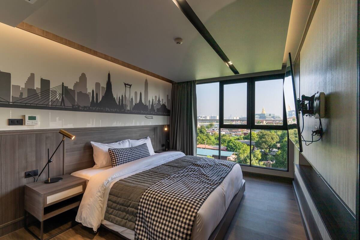 Quarto de hotel moderno com uma cama grande, uma mesa de cabeceira, um mural com vista para o horizonte da cidade e uma janela ampla que oferece uma vista urbana.