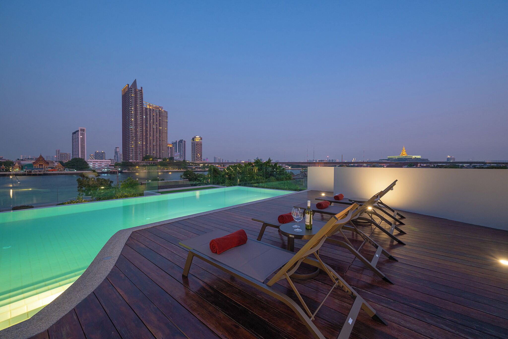 A rooftop pool with wooden deck and lounge chairs, overlooking a cityscape with high-rise buildings.