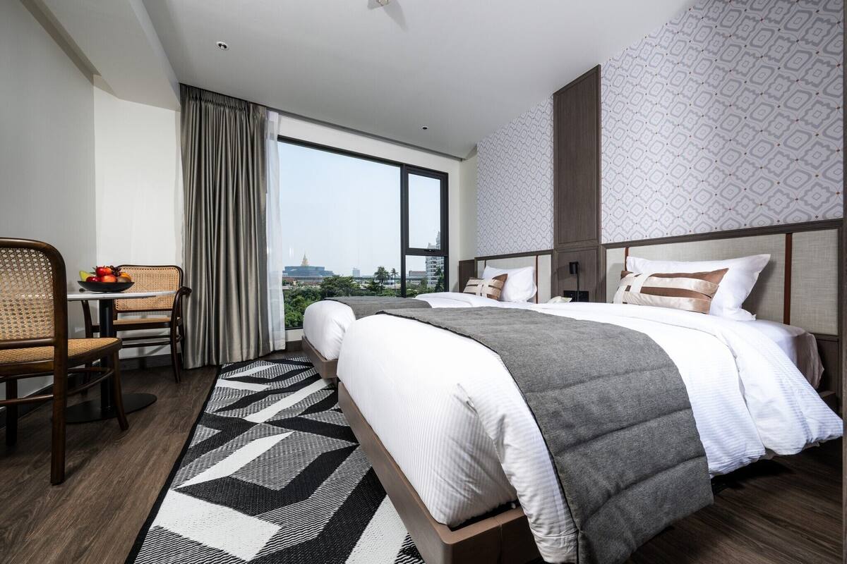 Quarto de hotel com uma cama grande, área de jantar com mesa e cadeiras, e vista para a cidade através de uma janela ampla.