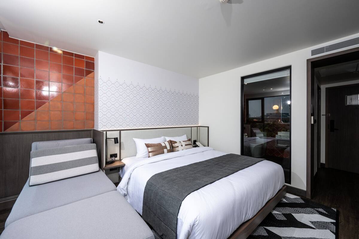 Quarto de hotel moderno com uma cama grande, um sofá, parede estampada e vista através de uma porta de vidro.