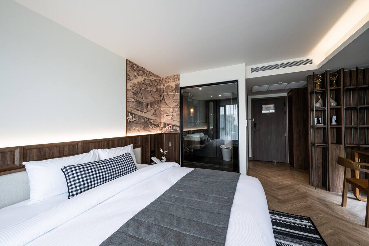 Quarto de hotel moderno com uma cama grande, cabeceira de madeira, uma obra de arte pendurada na parede, porta de vidro que dá para o banheiro e uma estante embutida.