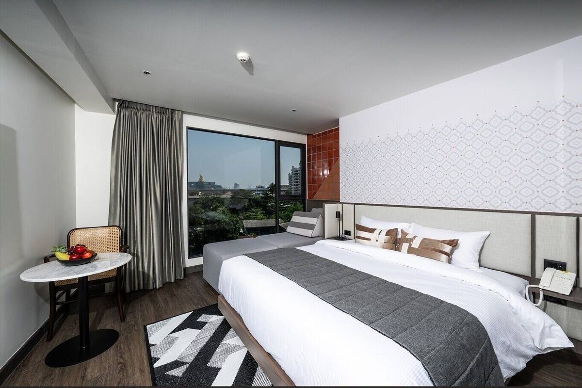 Quarto de hotel moderno com uma cama grande, uma mesinha com frutas, vista da cidade e parede estampada.