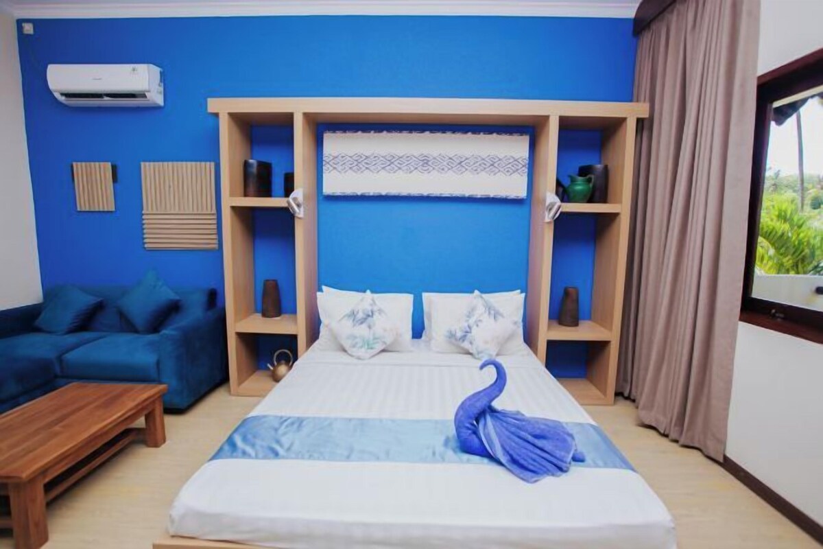 A bedroom with a bed, a blue wall, a window with curtains, a wooden shelf, and a small table.