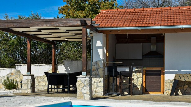 Un espace aménagé au bord de la piscine, comprenant un coin salon couvert, des murs en pierre et une cuisine avec un toit en tuiles.