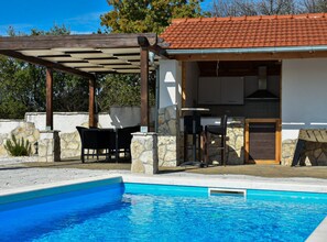 Un espace aménagé au bord de la piscine, comprenant un coin salon couvert, des murs en pierre et une cuisine avec un toit en tuiles.