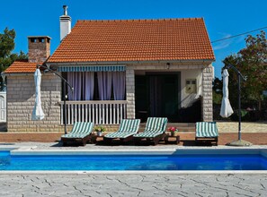 A poolside area with lounge chairs, a stone building with a tiled roof, and a clear blue pool.