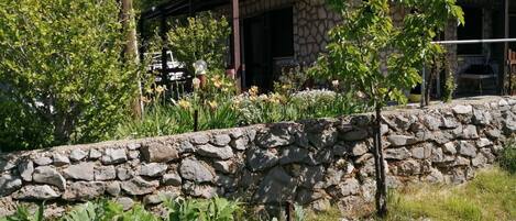 Una casa de piedra con un muro de piedra frontal, rodeada de vegetación y árboles.